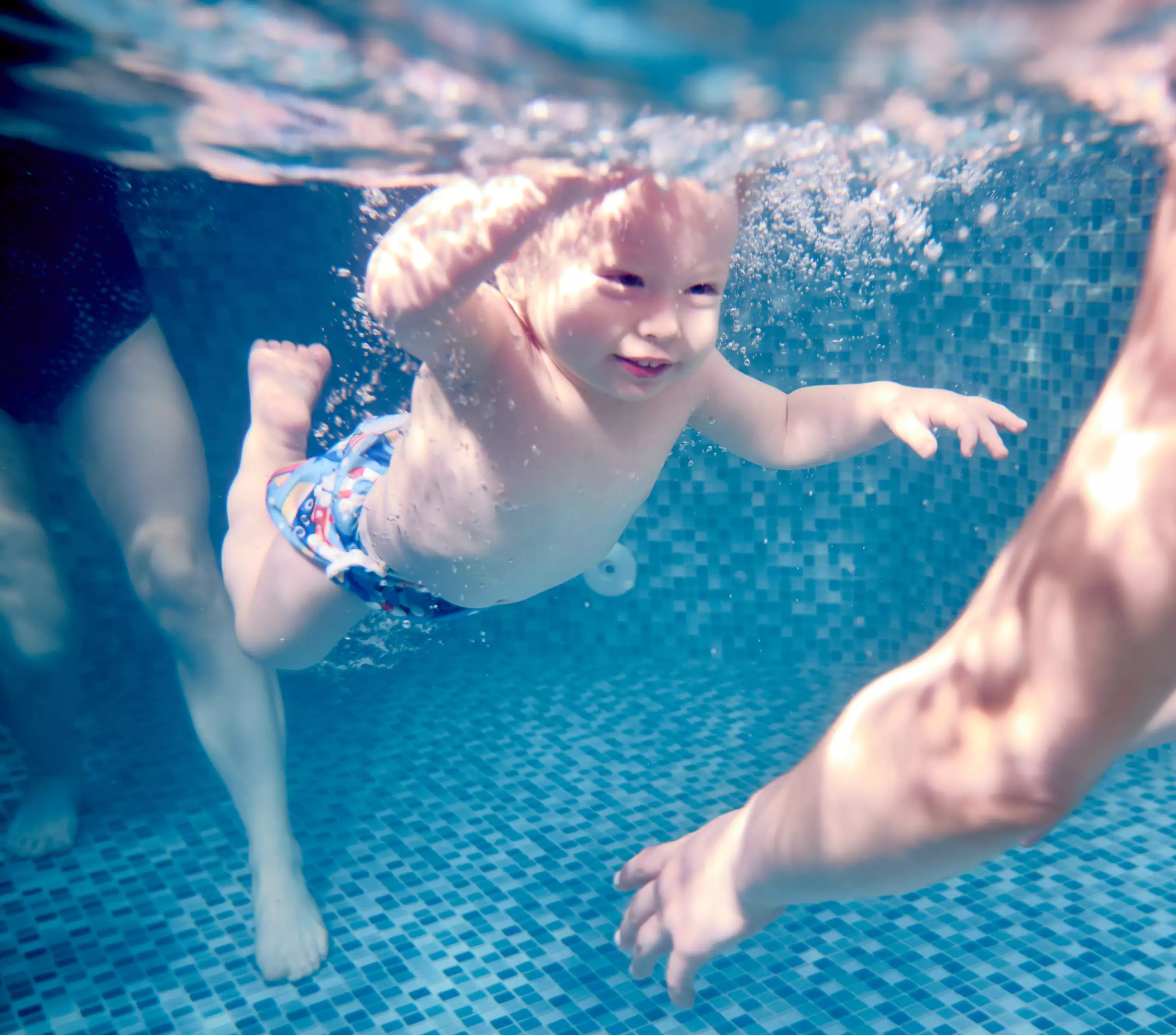 Kleines Kind taucht unter Wasser in einem Schwimmbecken mit blau-weiß gemusterten Fliesen, Erwachsene begleiten es.