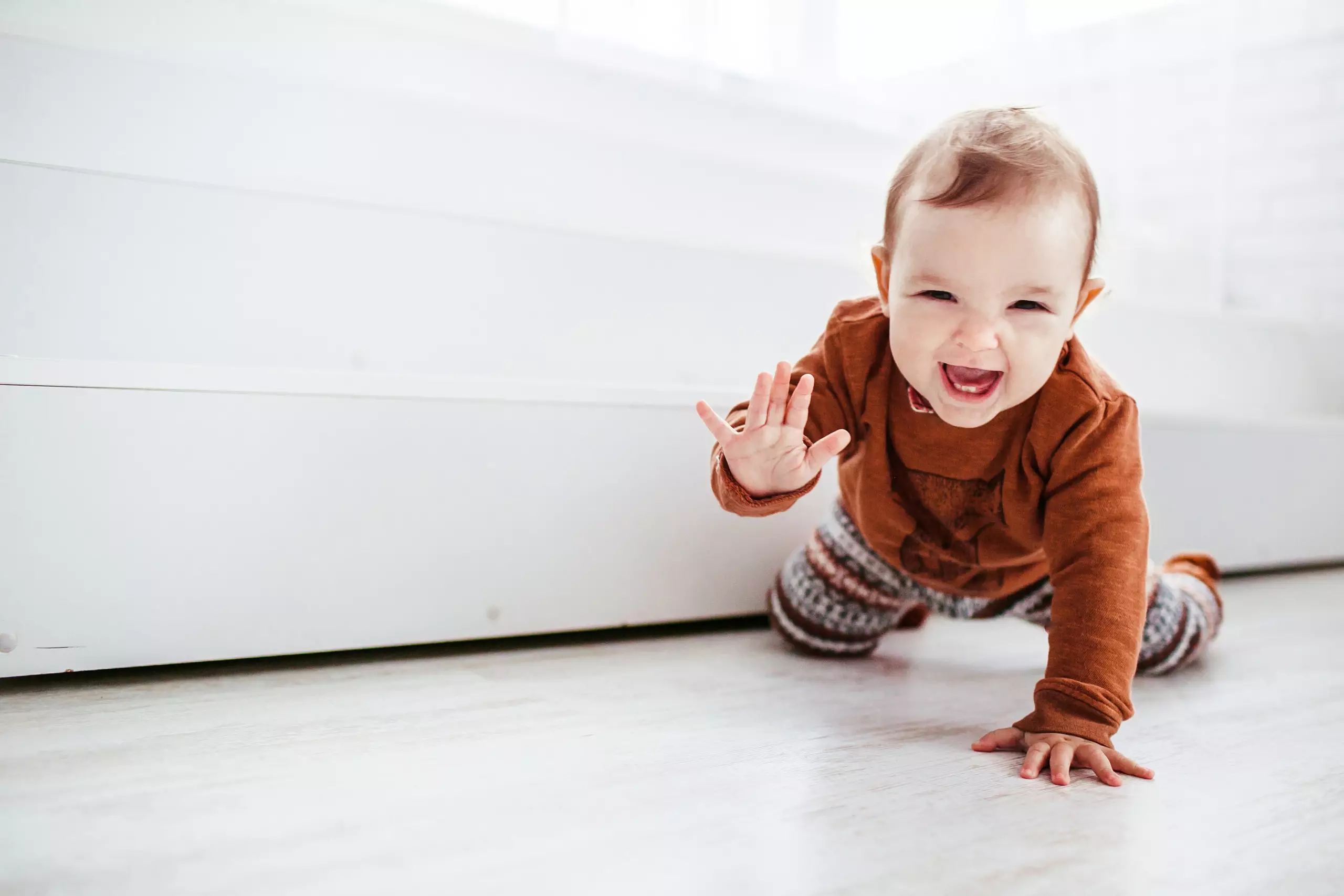 Lachendes Baby in braunem Pullover und gemusterten Hosen krabbelt auf hellem Boden und winkt mit der Hand