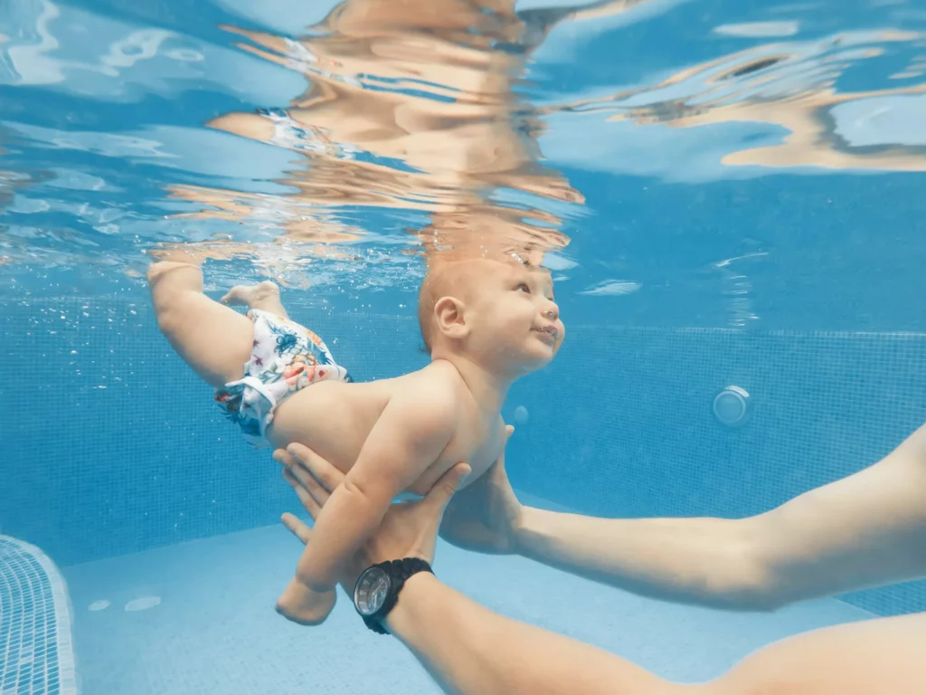 Baby mit Schwimmwindel wird unter Wasser in einem Pool von einer Person gehalten und lernt schwimmen