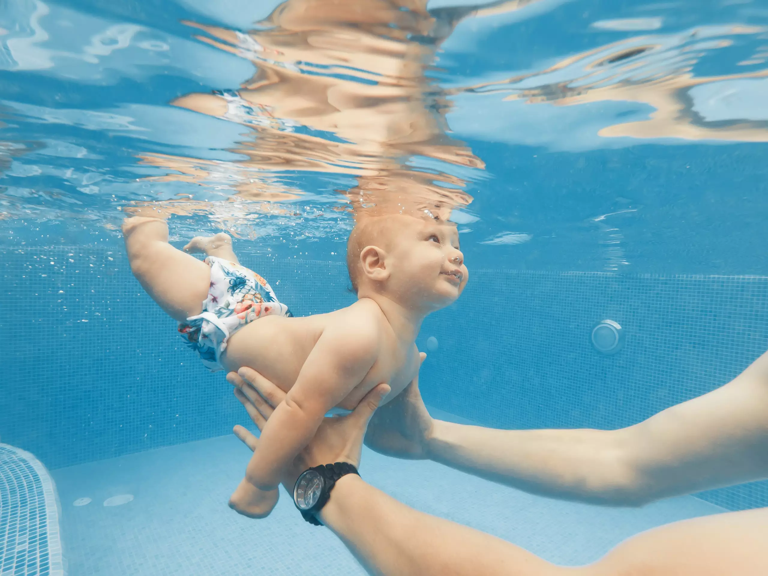 Baby mit Schwimmwindel wird unter Wasser in einem Pool von einer Person gehalten und lernt schwimmen