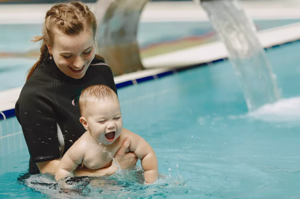 Frau mit Zopf hält lachendes Baby im Schwimmbecken mit Wasserfall im Hintergrund