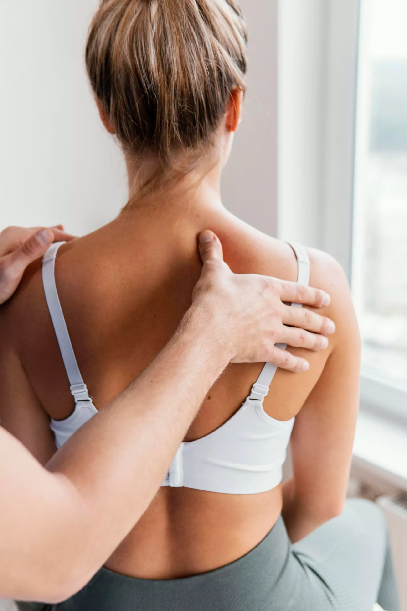 Frau in Sport-BH erhält Schulter- und Nackenmassage von Therapeut in hellem Raum mit Fensterlicht.