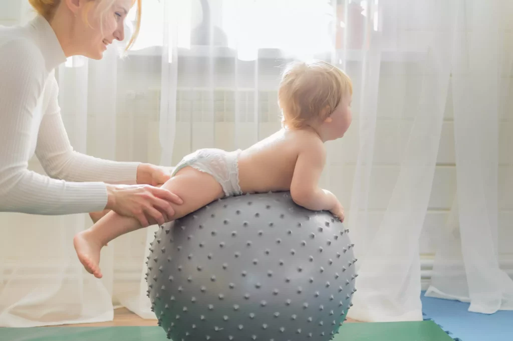 Kleines Kind liegt auf einem grauen Gymnastikball mit Noppen, Frau stützt es sanft beim Balancieren im hellen Raum