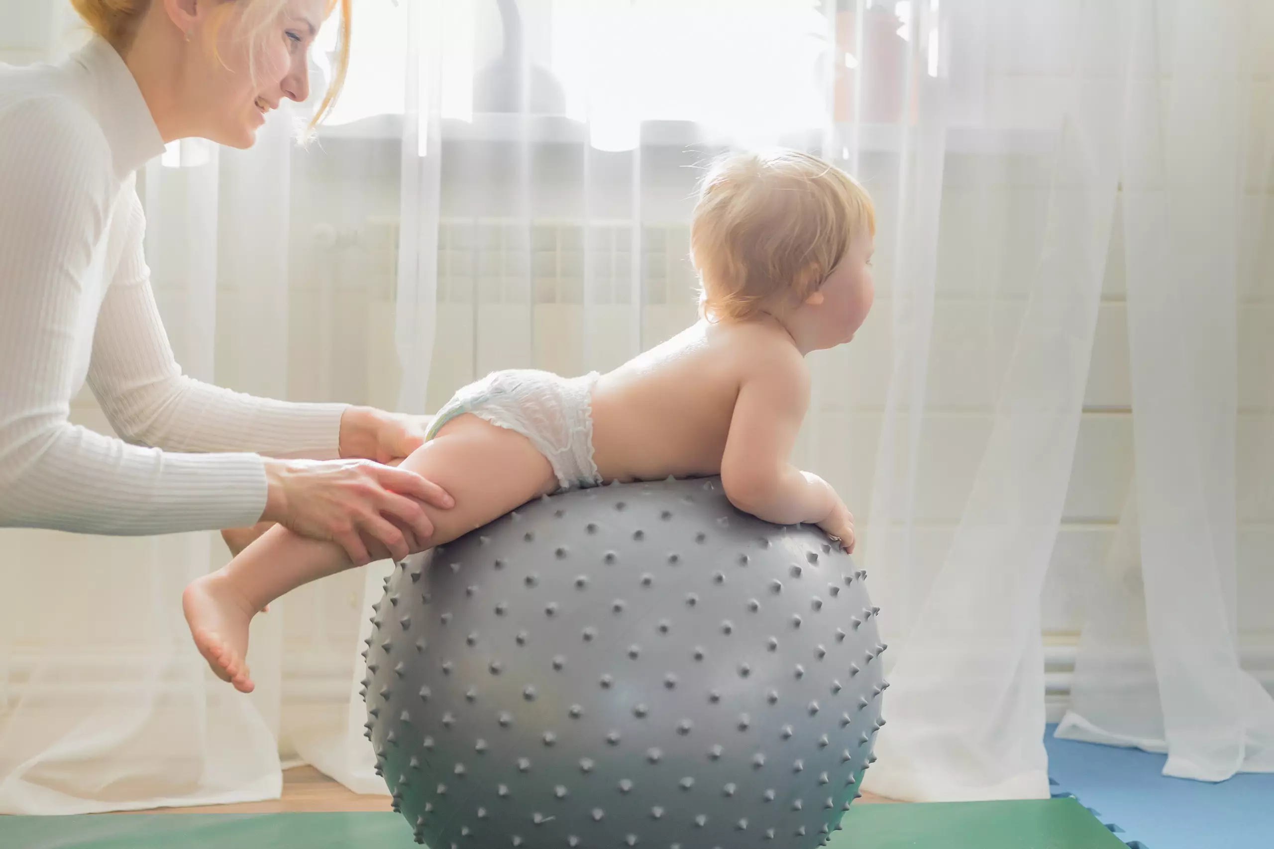 Kleines Kind liegt auf einem grauen Gymnastikball mit Noppen, Frau stützt es sanft beim Balancieren im hellen Raum